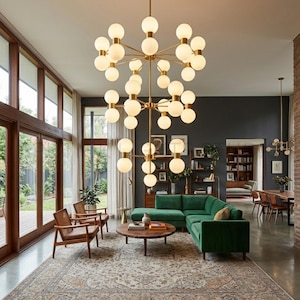 May include: A modern living room featuring a large, multi-bulb chandelier with a brass finish. The room includes a green sectional sofa, wooden chairs, a round coffee table, and a patterned rug. Large windows provide natural light.