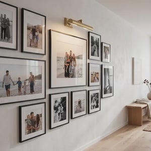 May include: A gallery wall arrangement featuring ten black picture frames of varying sizes containing black and white family photographs, illuminated by a brass wall-mounted picture light fixture centered above the largest central frame.