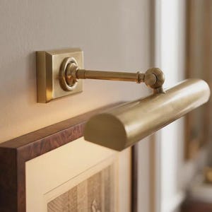 May include: A wall-mounted picture light with a brushed brass finish. The fixture features a square base plate, a horizontal cylindrical lamp head, and an adjustable arm with a pivot joint, positioned above a framed piece of artwork.