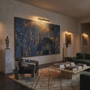 May include: A living room featuring a large three-panel abstract painting in deep blue and gold tones, a polished chrome armchair with black upholstery, a low square stone coffee table with books, and a dark green sofa on a patterned rug.
