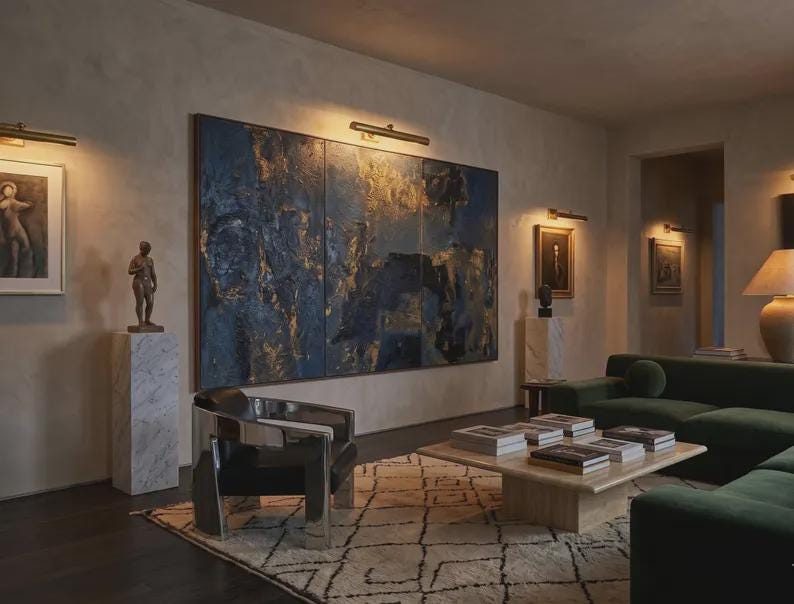 May include: A living room featuring a large three-panel abstract painting in deep blue and gold tones, a polished chrome armchair with black upholstery, a low square stone coffee table with books, and a dark green sofa on a patterned rug.