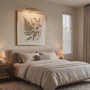 May include: A bedroom featuring a beige upholstered bed with a textured duvet, two wooden nightstands with small lamps, and a large framed botanical print of a fern and wildflowers hanging on the wall under a gold picture light fixture.