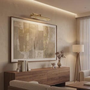 May include: A horizontal brass picture light fixture mounted above a large rectangular framed abstract painting with neutral beige and gray tones, positioned over a wooden sideboard cabinet with books and decorative ceramic objects.
