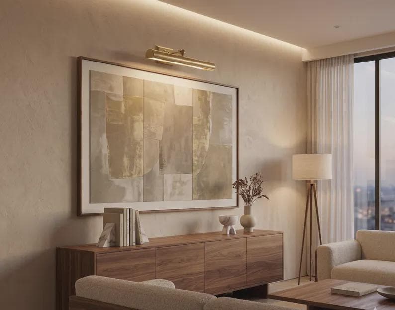 May include: A horizontal brass picture light fixture mounted above a large rectangular framed abstract painting with neutral beige and gray tones, positioned over a wooden sideboard cabinet with books and decorative ceramic objects.