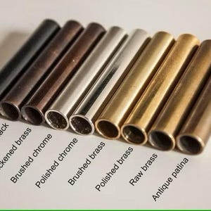 May include: A collection of nine cylindrical metal tubes arranged in a row, showcasing various finishes including matte black, antique black, blackened brass, brushed chrome, polished chrome, brushed brass, polished brass, raw brass, and antique patina.