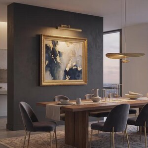 May include: A dining room featuring a rectangular wooden table with a light runner, surrounded by six dark gray upholstered chairs with gold legs. A large framed abstract painting with blue, white, and gold tones hangs on a dark wall.
