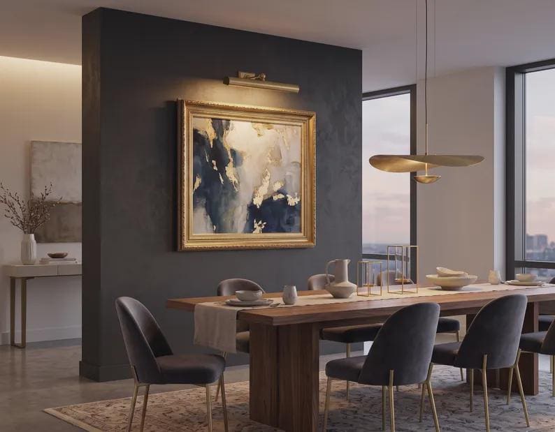 May include: A dining room featuring a rectangular wooden table with a light runner, surrounded by six dark gray upholstered chairs with gold legs. A large framed abstract painting with blue, white, and gold tones hangs on a dark wall.