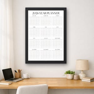 May include: A black framed 2026 Year Planner with a white background, displayed on a wooden desk. The planner is a grid layout with months and dates. A laptop, coffee cup, and desk lamp are also visible.