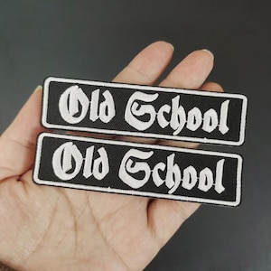 May include: Two black rectangular patches with the words "Old School" embroidered in white gothic lettering. The patches are rectangular with a white border and are likely for clothing or accessories.