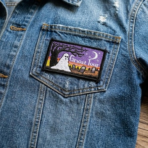 May include: Blue denim jacket with a rectangular patch on the pocket. The patch features a white ghost, a tree, and the words "Ghost Tour" against a purple sky. The jacket has a distressed look.