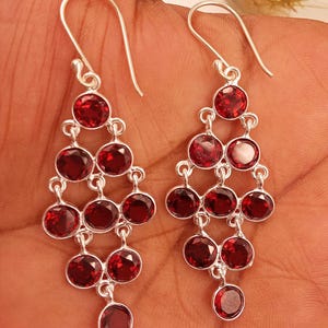 May include: A pair of silver chandelier earrings with multiple tiers of round, deep red gemstones. The earrings have a cascading design, with each tier connected by small silver links. The gemstones are a vibrant, polished red.