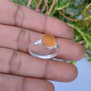 Op de afbeelding: Een zilveren ring met een gladde, afgerond vierkante oranje zeeglassteen in een gepolijste metalen zetting. De ringband is dun en van metaal, met een eenvoudig ontwerp dat de doorschijnende, levendige oranje edelsteen in het midden benadrukt.