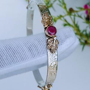 Può includere: Un bracciale rigido in argento con un intricato motivo in rilievo su tutta la fascia. Il centro è decorato con una pietra preziosa rotonda sfaccettata di colore rosa intenso, affiancata da due piccoli motivi a foglia in metallo dorato.