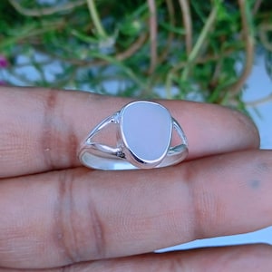 Könnte beinhalten: Ein Silberring mit einem glatten, undurchsichtigen, blassrosa, tropfenförmigen Stein, der in einer polierten Metallfassung sitzt. Die Ringschiene hat ein geteiltes Design, das an den Seiten der Steinfassung ansetzt und für einen schlichten Look sorgt.