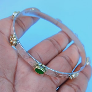 Pode incluir: Uma pulseira rígida de tom prateado com textura martelada. A banda é decorada com motivos florais dourados alternados e uma única pedra verde oval engastada. A superfície metálica tem um acabamento reflexivo em todo o design.