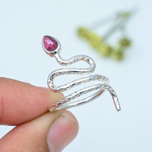 May include: A silver snake ring featuring a pink gemstone head. The ring coils around the finger, with textured detailing along the body. The gemstone is a vivid pink colour. The ring is likely made of silver.