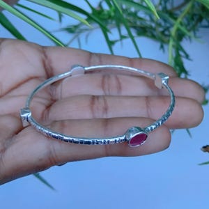 Puede incluir: Una pulsera rígida de plata con una superficie texturizada y martillada que presenta tres engastes distintos. Un engaste contiene una piedra preciosa roja lisa de forma ovalada, mientras que los otros dos parecen vacíos, mostrando el metal pulido.