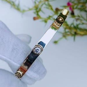 Puede incluir: Un brazalete rígido de tono plateado pulido con una piedra preciosa redonda y transparente en el centro engastada en un bisel dorado, acentuado por dos pequeños motivos circulares decorativos dorados con líneas radiantes.