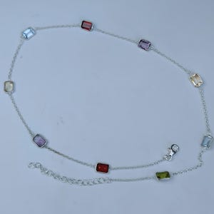 May include: A silver chain anklet featuring multiple rectangular, multi-coloured gemstones set in silver bezels. The stones include shades of red, purple, blue, green, and yellow. The anklet includes a lobster claw clasp and an adjustable chain link extension.