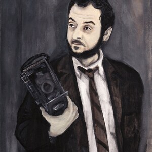 May include: A man in a dark suit and tie holds a vintage camera in his right hand. He is looking off to the side, and his expression is serious. The background is a dark gray.