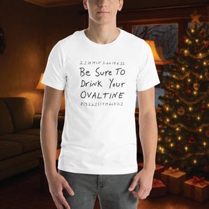 以下が含まれることがあります： 「Be Sure To Drink Your Ovaltine」の文字が黒い手書きフォントで書かれた白いTシャツ。テキストの上には数字、下にはさらに数字が書かれています。Tシャツはクリスマスツリーのある部屋で着用されています。