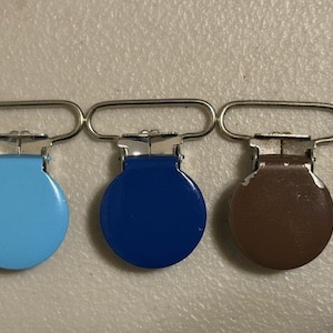 Puede incluir: Tres clips metálicos para chupete con diseño circular. Los clips tienen un lazo plateado y una base redonda y plana en azul claro, azul oscuro y marrón. Los clips están dispuestos en fila.