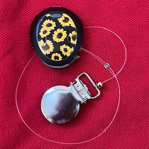 May include: A black pacifier clip with a sunflower design and a silver-colored metal clip. The clip is connected to the sunflower design by a clear, thin cord. The background is a solid red color.