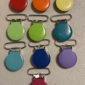 Pode incluir: Uma coleção de nove clipes de chupeta coloridos com fechos de metal. Os clipes apresentam uma variedade de cores brilhantes, incluindo vermelho, laranja, amarelo, verde, azul e roxo. Os clipes estão dispostos em uma superfície clara.