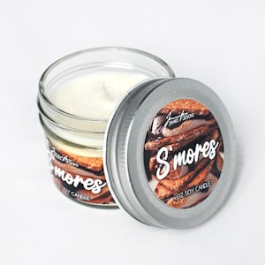 May include: A small glass jar candle with a silver lid. The label on the lid features a photo of a s'mores dessert and the text "S'mores 4 oz Soy Candle".