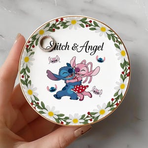 May include: A white ceramic ring dish with a gold rim, featuring a floral border of white daisies and red berries. The center displays a cartoon image of Stitch and Angel embracing, with the text "Stitch & Angel". A silver ring rests on the dish.