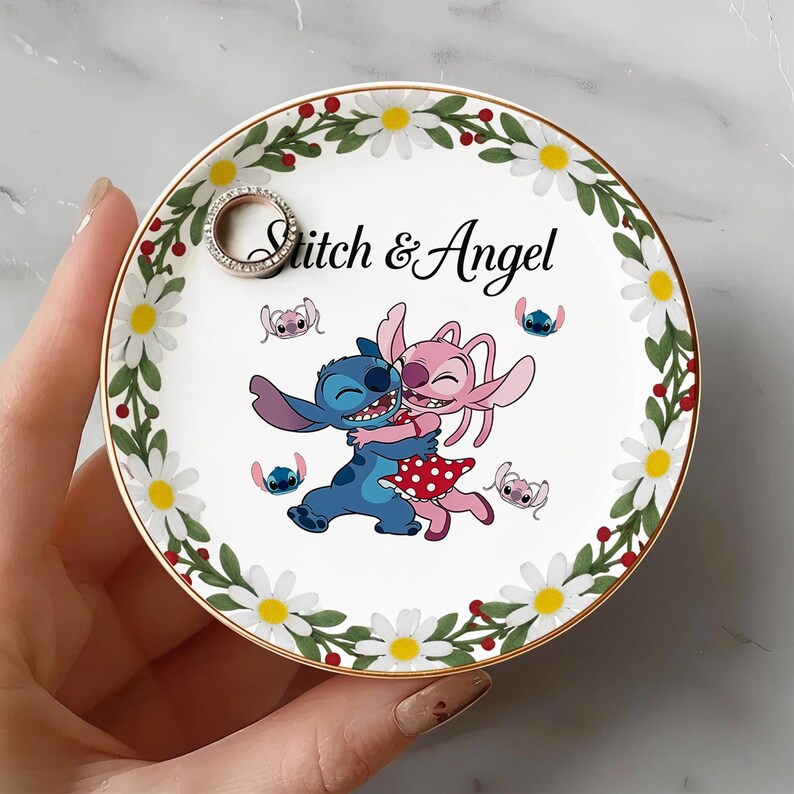 May include: A white ceramic ring dish with a gold rim, featuring a floral border of white daisies and red berries. The center displays a cartoon image of Stitch and Angel embracing, with the text "Stitch & Angel". A silver ring rests on the dish.