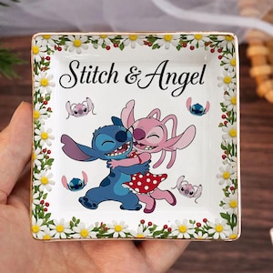 May include: Square ceramic trinket dish featuring a colorful illustration of Stitch and Angel embracing, with the text "Stitch & Angel". The dish is bordered with a floral design of white daisies and green leaves, with a gold trim.