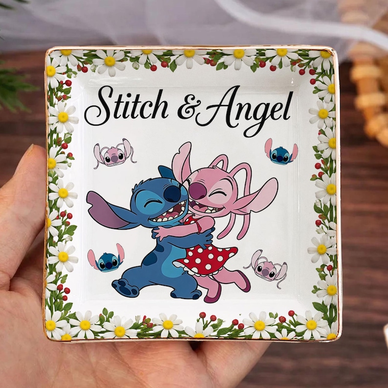 May include: Square ceramic trinket dish featuring a colorful illustration of Stitch and Angel embracing, with the text "Stitch & Angel". The dish is bordered with a floral design of white daisies and green leaves, with a gold trim.