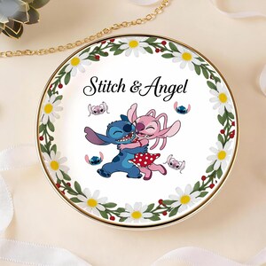 May include: A round decorative plate featuring the characters Stitch and Angel embracing, with the text "Stitch & Angel". The plate has a white center with a floral border of daisies and green leaves, and a gold rim.