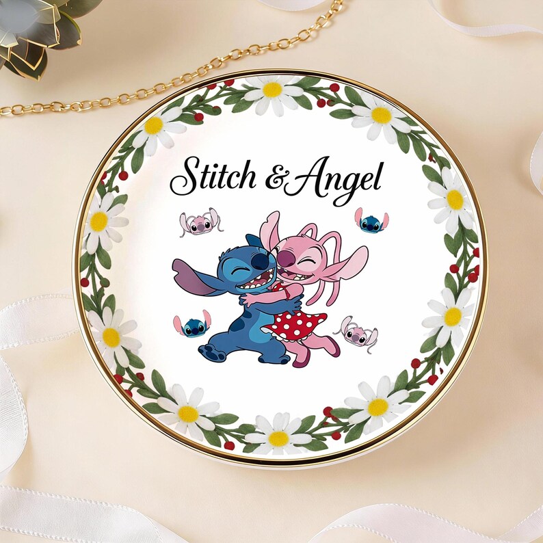 May include: A round decorative plate featuring the characters Stitch and Angel embracing, with the text "Stitch & Angel". The plate has a white center with a floral border of daisies and green leaves, and a gold rim.