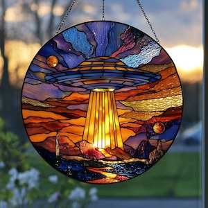 Può includere: Pannello rotondo in vetro colorato raffigurante un UFO sopra un paesaggio. L'opera presenta una combinazione di colori blu e arancione con un design dettagliato di un disco volante, raggi di luce e corpi celesti. Il pannello è sospeso da una catena.