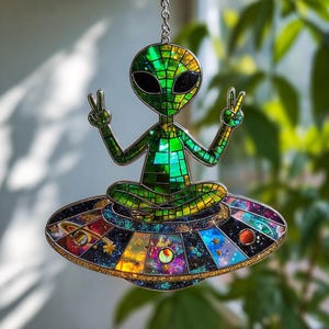 Può includere: Una figura aliena in vetro colorato in posizione seduta, con le braccia alzate a fare il segno della pace. L'alieno è verde con occhi neri, seduto su un'astronave circolare colorata. L'astronave presenta un design galattico con pianeti e stelle.
