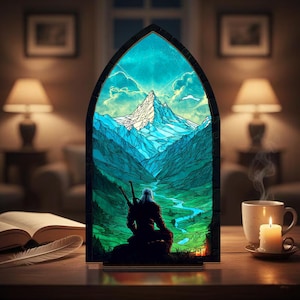 Może przedstawiać: Witryna ze szkła barwionego przedstawiająca górski krajobraz z sylwetką. Panel ma odcienie niebieskiego, zielonego i białego, w ciemnej, łukowatej ramie. Filiżanka kawy i zapalona świeca stoją na drewnianym stole.