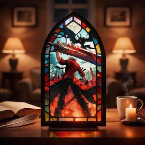 May include: Stained glass panel depicting a figure in a red coat with a sword, set against a fiery backdrop. The panel has a black frame and is on a wooden surface. A lit candle and a cup of coffee are placed nearby.