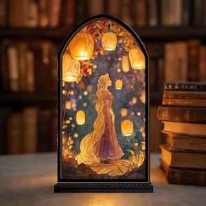 Può includere: Pannello decorativo in vetro colorato raffigurante una donna con lunghi capelli biondi in un abito viola, circondata da lanterne fluttuanti. Il pannello è incorniciato in una cornice ad arco nera con una base nera. Lo sfondo mostra un cielo stellato e un fogliame autunnale.