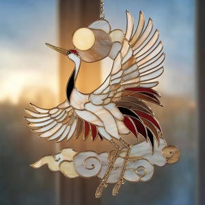 Könnte beinhalten: Glasfenster-Suncatcher mit einer Kranichfigur mit ausgebreiteten Flügeln, der auf einer Wolke sitzt. Der Vogel hat weißes, braunes und rotes Glas mit goldfarbenen Metallakzenten. Über dem Kranich befindet sich ein kreisförmiges Element, das die Sonne darstellt.