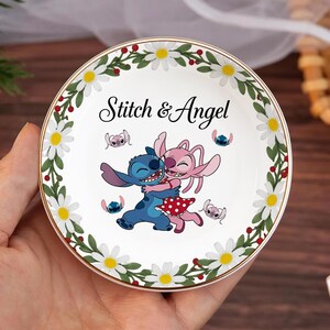 May include: A white ceramic trinket dish with a gold rim, featuring a floral border of daisies and green leaves. The center displays the words "Stitch & Angel" above an illustration of the characters Stitch and Angel embracing. Additional Stitch and Angel images are also present.