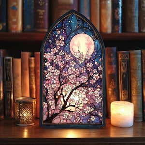 May include: A decorative stained glass lamp in the shape of a window, depicting a cherry blossom tree under a full moon. The lamp has a dark blue frame and is lit, showing pink and white flowers against a starry night sky. A candle and a small lantern are also present.