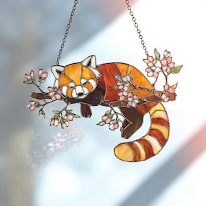 May include: Stained glass red panda suncatcher. The artwork features a red panda resting on a branch with pink blossoms. The suncatcher is made with shades of orange, brown, and white, and is suspended by a metal chain.