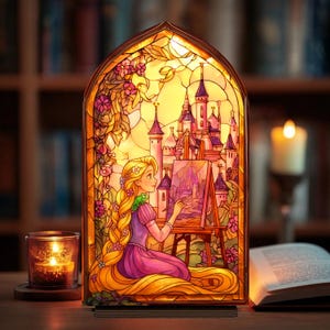 Peut inclure: Lampe en vitrail représentant une princesse peignant un château. La lampe dégage une lumière chaleureuse, avec une illustration détaillée d'un château, de fleurs et d'une princesse aux longs cheveux blonds. Une bougie et un livre ouvert sont au premier plan.