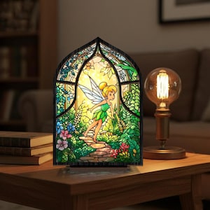 Könnte beinhalten: Buntglaslampe mit einer Fee in einer Gartenszene. Die Lampe hat einen dunklen Holzrahmen und einen warmen, gelben Schein. Die Fee ist in Grün und Gelb dargestellt, umgeben von bunten Blumen und Blättern.