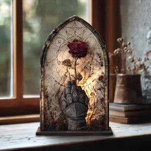 May include: A gothic-style decorative lamp featuring a red rose held by a metallic gauntlet. The lamp has an arched frame with an aged parchment-like background, and is set on a wooden base. The design incorporates floral and geometric elements.