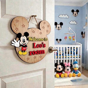 Könnte beinhalten: Ein helles Holz-Türschild in Form von Mickey-Mouse-Ohren mit einer Mickey-Mouse-Grafik und dem Text "Welcome to Leo's room" in Rot und Gelb. Das Schild ist an einem braunen Seil aufgehängt und mit eingravierten Sternumrissen verziert.