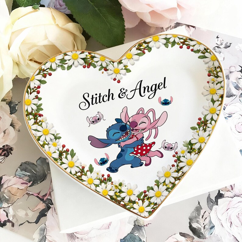 May include: Heart-shaped decorative plate with a gold rim, featuring the characters Stitch and Angel embracing. The plate is white with a floral border of daisies and red berries. The text "Stitch & Angel" is written in black script.