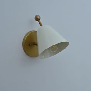 May include: A white cone-shaped wall sconce with a brass-colored base and arm. The light fixture has a small, round brass accent at the top. The sconce is designed to be mounted on a wall and provides focused lighting.